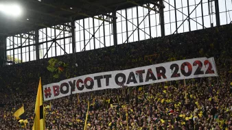 L'image montre un grand groupe de supporters dans un stade, portant des vêtements jaunes et noirs. Ils tiennent une immense banderole sur laquelle est inscrit "#BOYCOTT QATAR 2022". L'atmosphère semble dynamique et engagée, avec un fort sentiment de solidarité parmi les fans. Les tribunes sont pleines, créant une ambiance électrisante.