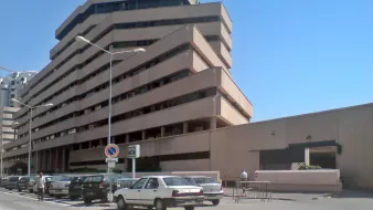 L'image montre un bâtiment moderne à plusieurs étages avec une architecture angulaire et des balcons en saillie. La façade est en béton, avec des couleurs neutres. On peut voir des voitures garées le long de la route, et il semble y avoir des piétons dans la zone. Le ciel est dégagé, ce qui donne une atmosphère ensoleillée à la scène. Le bâtiment dégage une impression de fonctionnalité et de modernité.