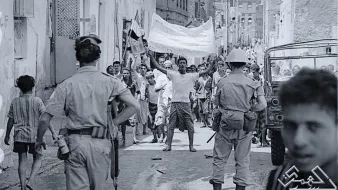 L'image montre une scène de rue dans un contexte de manifestation ou de rassemblement. On peut voir des groupes de personnes, certains brandissant des drapeaux ou des pancartes, exprimant visiblement leur émotion ou leur revendication. Des policiers réglementent la situation, et l'atmosphère semble tendue. L'environnement urbain est composé de bâtiments simples, typiques d'une ville. Les vêtements des gens indiquent peut-être une époque passée, soulignant le contexte historique de la scène.