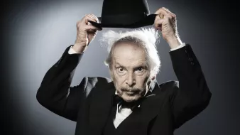 L'image montre un homme âgé portant un costume noir avec un nœud papillon. Il tient un chapeau melon avec une main, et son autre main semble légèrement levée. Ses cheveux sont ébouriffés, ce qui donne un aspect excentrique à son expression. L'arrière-plan est sobre, avec une lumière qui crée un effet dramatique, mettant en valeur son visage et son chapeau.