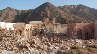 L'image montre un site de ruines où les structures sont partiellement effondrées, avec des débris éparpillés sur le sol. Des échafaudages sont visibles autour des bâtiments, suggérant un effort de restauration ou de démolition. En arrière-plan, on aperçoit une montagne aux pentes douces, recouverte de verdure, sous un ciel dégagé. L'ensemble dégage une impression de désolation mêlée à la beauté naturelle environnante.