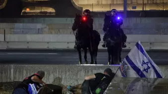 L'image montre une scène de tension en milieu urbain, avec deux policiers à cheval qui surveillent la situation. En bas, on aperçoit des manifestants, dont certains semblent se protéger ou être en détresse. Un drapeau israélien est visible au sol, ajoutant une dimension politique à la scène. L'éclairage des policiers et l'atmosphère générale suggèrent un climat de conflit ou de protestation.