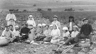 L'image montre un groupe de personnes rassemblées en pleine nature, probablement dans une zone agricole. Les individus, habillés de vêtements traditionnels de l'époque, semblent se préparer à un repas ou à une pause. Certains tiennent des objets comme des paniers et des outils agricoles. Le paysage en arrière-plan est rural, avec des champs et des collines. L'atmosphère dégage une impression de communauté et de travail collectif.