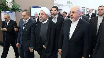 این تصویر گروهی از افراد را نشان می‌دهد که در حال حرکت به سمت یک مکان هستند. یکی از آنها به وضوح لباس روحانیت پوشیده و دیگران نیز به طور رسمی لباس پوشیده‌اند. به نظر می‌رسد این افراد در یک رویداد یا کنفرانس رسمی شرکت کرده‌اند. محیط تصویر ممکن است یک مکان اداری یا کنفرانس باشد که نشان‌دهنده تعاملات سیاسی یا اقتصادی است.