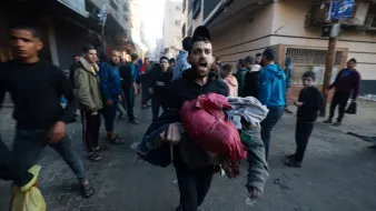 L'image montre une scène urbaine animée où un homme, visiblement en détresse, transporte des vêtements ou des objets dans ses bras. Il semble crier ou exprimer une émotion forte, tandis que d'autres personnes marchent autour de lui. L'environnement est urbain, avec des bâtiments en arrière-plan qui semblent endommagés ou en délabrement. L'atmosphère de l'image suggère une situation de crise ou d'urgence, avec une foule qui se déplace rapidement.