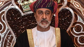 L'image montre un homme assis sur un trône orné. Il porte un costume traditionnel, comprenant une robe blanche et un turban coloré. Son expression est sérieuse et solennelle. Le fond est richement décoré avec des motifs complexes, créant une ambiance majestueuse. Les détails de la couronne et du trône mettent en valeur son statut élevé.