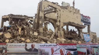 تظهر الصورة مشهداً لمدينة مدمرة، حيث يمكن رؤية مبانٍ مهدمة مع حطام في كل مكان. يوجد فيها جنود مع وجود لافتات تحمل شعارات سياسية وصور لشخصيات مؤثرة. الخلفية تعكس آثار الحرب والدمار، بينما تعبر اللافتات عن مواقف سياسية معينة. الأجواء تعكس حالة من التوتر والصراع.