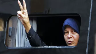 L'image montre une femme qui fait un signe de paix avec sa main, en levant deux doigts. Elle est visible à travers une fenêtre légèrement ouverte d'un véhicule. La femme porte un hijab. Son expression semble déterminée et engagée. Le fond est sombre, ce qui met en avant son geste et son visage.