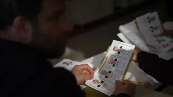 En la imagen se puede ver a una persona sosteniendo una boleta electoral. La boleta muestra varias fotografías de candidatos, junto con sus nombres y posiblemente posiciones a las que aspiran. La persona parece estar concentrada en la boleta, mientras que en la mesa hay más documentos. La escena parece tener lugar en un ambiente de votación.