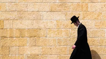 L'image montre un homme marchant le long d'un mur de pierre de couleur beige. Il est habillé d'un long manteau noir et porte un chapeau noir. Son expression est sérieuse et il semble concentré sur son chemin. L'arrière-plan est uni, ce qui met en valeur la silhouette de l'homme qui avance seul.