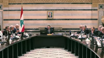 تصویر یک جلسه رسمی دولتی در لبنان را نشان می‌دهد. تعدادی از مقامات و اعضای دولت در اطراف یک میز بزرگ نشسته‌اند. در مرکز جلسه، یک مرد با لباسی رسمی و آرامش بر روی صندلی ریاست نشسته است و به دیگران نگاه می‌کند. در پس‌زمینه، پرچم لبنان و تصویری از یک شخصیت مهم بر دیوار نصب شده است. فضای اتاق با خطوط عمودی و افقی تزئینی و رنگ‌های ملایم طراحی شده است. افراد حاضر در جلسه به موضوعات مختلفی مشغول بحث و تبادل نظر هستند.