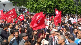 L'image montre une foule rassemblée dans une rue, lors d'une manifestation. Les participants tiennent des drapeaux rouges, probablement portant des symboles ou des slogans liés à la Tunisie. Il y a une grande variété de personnes dans la foule, de différents âges et expressions. L'ambiance semble énergique, avec des signes d'engagement et de participation active. En arrière-plan, on peut voir des bâtiments et des arbres, ce qui indique que l'événement se déroule dans un endroit urbain.