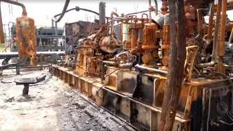 L'image montre une installation industrielle endommagée, probablement suite à un incendie. On peut voir des équipements métalliques, tels que des machines et des tuyaux, recouverts de rouille et de suie. Le sol est noirci par des résidus, indiquant les conséquences d'un sinistre. L'environnement semble très dégradé, avec des éléments de construction en arrière-plan également affectés par des dommages. L'ensemble donne une impression de désolation et de négligence.