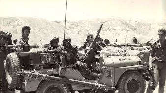 L'image montre un groupe de soldats réunis autour d'un véhicule militaire, probablement une jeep. Ils semblent engagés dans une mission, avec des équipements militaires visibles sur le véhicule. Le paysage en arrière-plan est aride, avec des collines ou des montagnes. Les soldats prennent la pose, certains souriant, et semblent confiants dans leur environnement. L'atmosphère dégage un sentiment de camaraderie et de détermination.