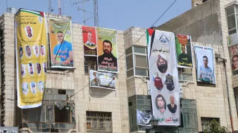 L'image montre un bâtiment avec plusieurs affiches et bannières accrochées sur sa façade. Ces affiches présentent des portraits de différentes personnes, probablement des figures politiques ou des militants, accompagnées de textes en arabe. Les couleurs des bannières sont vives, avec du jaune, du vert et du rouge, et beaucoup d'entre elles semblent commémorer ou soutenir des individus spécifiques. On peut également voir des drapeaux et d'autres éléments décoratifs liés à des mouvements politiques. L'environnement urbain donne une impression d'animation et de vie locale.