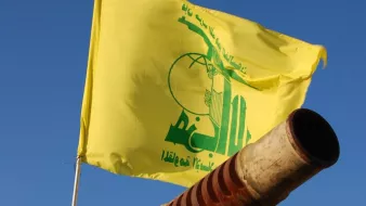L'image montre un canon pointant vers le ciel, avec un drapeau jaune flottant au-dessus. Sur le drapeau, il y a des inscriptions en arabe ainsi qu'un logo symbolique. Le fond est bleu, suggérant une journée ensoleillée. Le canon présente des signes d'usure, avec une surface rouillée.