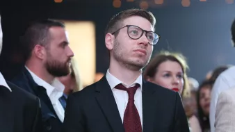 Nell'immagine si può vedere un gruppo di persone in un contesto che sembra essere un evento sociale o una festa. Un uomo in primo piano indossa un completo scuro con una cravatta bordeaux e occhiali. Ha un'espressione pensierosa, mentre osserva attentamente ciò che accade di fronte a lui. Dietro di lui ci sono altre persone, alcune delle quali sembrano anch'esse concentrate sull'eventuale spettacolo o presentazione. L'atmosfera è elegante e formale, con una leggera illuminazione che aggiunge un tocco di atmosfera.