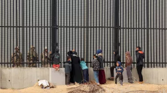 The image depicts a group of people, including women and children, standing in front of a tall, barred fence. On the other side of the fence are soldiers or guards, who appear to be watching the group. The scene conveys a sense of separation and tension, with barbed wire in the foreground emphasizing the barriers between the two sides. The individuals on the left seem to be engaging with the guards, possibly seeking assistance or communication. The overall atmosphere suggests a complex and challenging situation, likely related to migration or security issues.