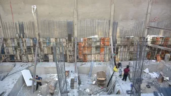 L'image montre un chantier de construction. On peut y voir des ouvriers qui travaillent dans une excavation, entourés de barres d'acier et de matériaux de construction. Les murs en béton sont partiellement visibles, avec des panneaux de coffrage en bois et en métal disposés verticalement. L'environnement semble être en cours de développement, avec une variété d'outils et de matériaux éparpillés autour de l'espace de travail.