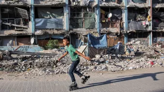 L'image montre un jeune garçon qui skates sur des rollers, se déplaçant avec agilité sur un trottoir. En arrière-plan, des bâtiments en mauvais état sont visibles, avec des fenêtres cassées et des débris éparpillés, ce qui évoque un environnement urbain dégradé. Des morceaux de tissu et d'autres objets sont accrochés aux façades, ajoutant à l'impression générale de désolation. Le contraste entre l'activité du garçon et le décor environnant souligne une certaine résilience face à la pauvreté.