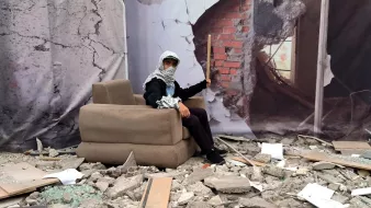 L'image montre une scène de destruction avec des débris éparpillés au sol, tels que des morceaux de béton et des morceaux de verre brisé. Au centre, une personne est assise sur un canapé apparemment plus ou moins intact. Elle porte un foulard autour de la tête et tient un bâton. En arrière-plan, il y a une illustration d'un mur brisé, ajoutant à l'impression de désolation et de chaos de la scène.