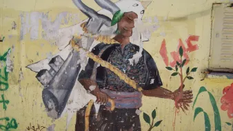 L'image montre une fresque sur un mur. On y voit un personnage portant un costume traditionnel, avec un air déterminé. Ce personnage tient une plante dans une main, symbolisant la vie et la nature. Sur son dos, on peut apercevoir une sorte d'outil ou d'équipement qui pourrait symboliser le travail ou un engagement envers la terre. Le fond est de couleur jaune, ce qui attire l'œil et met en avant le contraste avec les couleurs du personnage. Des inscriptions et des graffitis en arrière-plan ajoutent du caractère à l'œuvre.