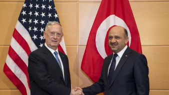 L'image montre deux hommes se serrant la main, probablement en signe de coopération ou d'accord. À l'arrière-plan, on peut voir deux drapeaux : celui des États-Unis à gauche et celui de la Turquie à droite. Les deux hommes portent des costumes formels, et l'ambiance semble être celle d'une rencontre officielle.