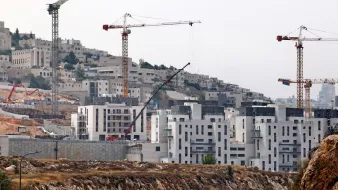 L'immagine mostra un cantiere edile situato in una zona collinare. Sono visibili diverse gru da costruzione, che si ergono sopra gli edifici in fase di completamento. Le strutture in costruzione hanno una forma moderna e presentano superfici di cemento. Sullo sfondo si intravedono altre costruzioni e il panorama collinare circostante, con vegetazione sparsa. L'atmosfera sembra essere quella di un'area in sviluppo e crescita urbana.