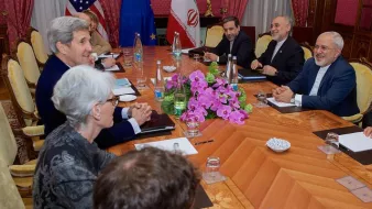 L'image montre une réunion diplomatique autour d'une grande table en bois. Des personnes sont assises de chaque côté, impliquées dans une discussion. On aperçoit des drapeaux d'États-Unis, de l'Union européenne et d'Iran sur la table. Au centre, il y a un bouquet de fleurs, probablement des orchidées. L'ambiance semble sérieuse mais productive, avec des participants au visage souriant, indiquant un échange positif.