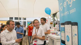 L'image montre une scène animée dans un événement, probablement un salon ou une foire. On peut voir des personnes interagissant autour d'un stand. Un homme, habillé en chemise blanche, semble discuter avec une jeune femme qui porte un uniforme avec un logo. À l'arrière-plan, il y a une bannière portant le logo d'Engie et des représentations d'applications sur des smartphones. L'ambiance est conviviale, avec des gens qui semblent engagés et intéressés par ce qui se passe.
