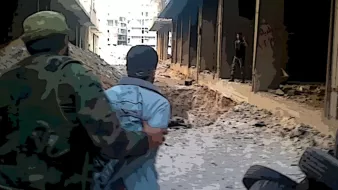 L'image montre une scène de guerre urbaine où deux individus se trouvent dans un environnement dévasté. On peut voir un soldat en uniforme qui tient une personne par l'épaule, tandis qu'un autre homme est visible au loin, apparemment dans une position de menace. Le sol est rempli de décombres, et l'atmosphère générale semble tendue et dangereuse, illustrant les ravages d'un conflit armé. Les bâtiments environnants sont endommagés et abandonnés.
