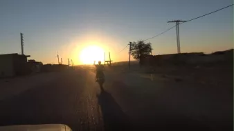 The image depicts a scenic view of a road extending into the distance during sunset. A person on a motorcycle is riding away from the camera, with their hand raised, possibly in a wave or gesture. The sun is setting on the horizon, casting a warm golden light across the landscape. On either side of the road, there are silhouettes of buildings and trees, and the scene conveys a sense of tranquility and adventure. Utility poles line the road, contributing to the rural setting.