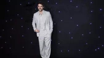 L'immagine mostra un uomo in piedi su uno sfondo scuro, illuminato da piccole luci bianche che brillano. Indossa un completo grigio chiaro, caratterizzato da pantaloni ampi e una giacca ben tagliata. La sua camicia è di un tono chiaro e ha il colletto aperto. L'uomo ha un sorriso e porta dei baffi, dando un'aria elegante e sicura di sé.