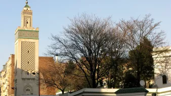 L'image montre une vue d'un bâtiment architectural distinctif, probablement une mosquée, avec un minaret élancé décoré de motifs. En arrière-plan, on aperçoit des arbres dépouillés, ce qui indique peut-être une saison hivernale ou automnale. Le mur entourant le bâtiment est blanc avec des bordures vertes. Des personnes marchent le long de la rue, créant une atmosphère de vie urbaine. Le ciel est dégagé, suggérant une journée ensoleillée.