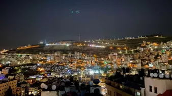The image shows a nighttime view of a cityscape, likely on the slope of a hill or mountain. The scene is illuminated with numerous lights from buildings, giving it a vibrant and lively atmosphere. In the foreground, there are rooftops with antennas or satellite dishes visible, while the background features the illuminated hillside, possibly with telecommunications towers or other structures visible at the top. The sky is dark, suggesting it's a clear night with some stars or light reflections. Overall, the image conveys a sense of urban life and a stunning nocturnal landscape.