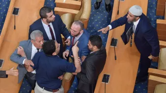 The image shows a heated confrontation occurring in a legislative assembly. Several men are engaged in a tense discussion or argument, with some gesturing animatedly. The setting appears to be a formal chamber, indicated by the rows of seats and the official appearance of the individuals. Some men are standing closely together, while others seem to be pointing or raising their hands, indicating strong emotions. Overall, the scene conveys a moment of conflict or debate among the legislators.