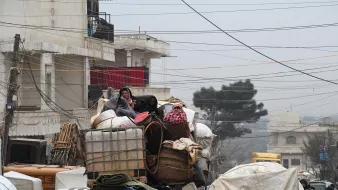 در این تصویر، گروهی از افراد در حال سفر بر روی یک کامیون بار هستند. آن‌ها به طور واضحی در میان بار زیادی از محصولات و وسایل قرار دارند. برخی از افراد پوشیده از لباس‌های ضخیم و شال‌های رنگی به نظر می‌رسند که به دلیل سرما به خود پیچیده‌اند. محیط اطراف شلوغ و شهرنشین به نظر می‌رسد، با ساختمان‌ها و خطوط برق مشخص. وضعیت کلی تصویر احساس چالش و سختی را القا می‌کند.
