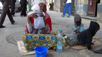 تظهر في الصورة امرأة تجلس على الأرض، تحمل طفلًا في حضنها بينما يبدو أنها ترعى طفلين آخرين يجلسان بجانبها. المرأة ترتدي ملابس تقليدية ذات ألوان زاهية، ويظهر التعب على وجهها. الأطفال يبدون مشغولين بلعب أو بتفاعل مع البيئة المحيطة. في الخلفية، يمكن رؤية بعض المارة والمباني، مما يوحي بأن الصورة التقطت في منطقة حضرية. يوجد أيضًا زجاجة ماء ووعاء أزرق بالقرب من المرأة.