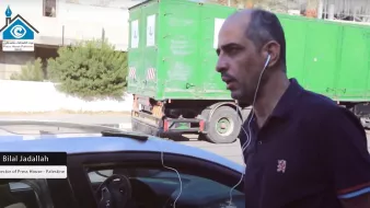 Nell'immagine si vede un uomo che cammina accanto a un'auto. Indossa una maglietta scura e ha delle cuffie che gli pendono dalle orecchie. Sullo sfondo si nota un camion verde e alcune piante. L'uomo sembra concentrato e diretto verso un punto specifico. A sinistra, è presente un'etichetta che indica il suo nome e la sua posizione, come direttore di un'organizzazione legata alla stampa in Palestina.