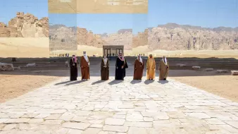 L'immagine mostra un gruppo di uomini vestiti con abiti tradizionali, dimostrando un'atmosfera di formalità e celebrazione. Sono disposti in fila su una pavimentazione di pietre chiare, con un paesaggio desertico sullo sfondo. Dietro di loro si erge una struttura riflettente che sembra integrarsi con l'ambiente circondante, creando un gioco di luci e riflessi. Le montagne in lontananza aggiungono profondità alla scena, conferendo un senso di grandiosità e atmosfera unica.