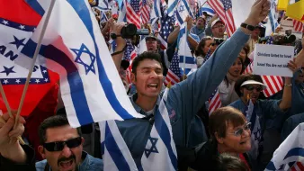 The image depicts a large gathering of people holding flags, primarily Israeli and American, as they participate in a rally or demonstration. The crowd appears to be energetic and passionate, with individuals expressing enthusiasm—some are cheering, while others are waving their flags. The setting seems to be outdoors, likely in a public space, and the atmosphere conveys a sense of unity and support for a cause related to Israel.