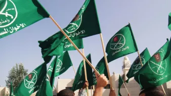 The image depicts a crowd of people holding up green flags. The flags feature a logo that includes Arabic text and a symbol, likely associated with a political or religious group. The scene appears to be one of a gathering or demonstration, with an emphasis on the flags being waved prominently. The background suggests an outdoor setting, possibly near a mosque or a public place.