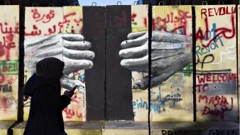 L'image montre un mur avec des graffitis colorés, dont le mot "Révolte" et d'autres messages en arabe et en anglais. Au centre, on peut voir des mains peintes qui semblent se toucher à travers une fissure du mur. Une silhouette d'une personne portant un hijab marche devant ce mur, avec un téléphone à la main. L'ensemble évoque des thèmes de résistance et de lutte sociale.
