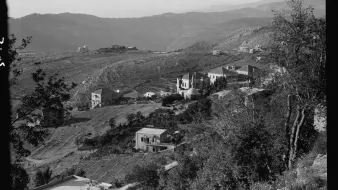 L'image montre un paysage montagneux avec des collines qui s'étendent à l'horizon. On peut apercevoir plusieurs maisons éparpillées sur les flancs des collines, entourées de végétation. La scène semble être prise en noir et blanc, ce qui lui donne un aspect nostalgique. Les terrains agricoles sont visibles, indiquant une zone rurale. Les montagnes en arrière-plan ajoutent une profondeur à la composition paysagère.
