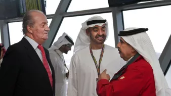 En la imagen se pueden ver a tres hombres conversando. Dos de ellos visten trajes tradicionales árabes, mientras que el tercero lleva un traje negro con una camisa blanca y una corbata roja. El entorno parece ser un aeropuerto o un lugar moderno. Los hombres están interactuando de manera amigable y hay pantallas en el fondo, lo que sugiere un ambiente ocupado y profesional.