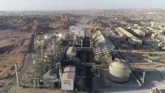 L'image présente une vue aérienne d'un complexe industriel. On peut observer plusieurs bâtiments de grande taille, comprenant des structures cylindriques et pyramidales. La zone environnante semble aride avec des collines et des bâtiments à l'arrière-plan. Des tuyaux et des infrastructures industrielles sont visibles, indiquant une activité de production. On discerne également de la vapeur s'échappant d'une des installations, suggérant un processus en cours. L'ensemble dénote un environnement industriel dans une région désertique.