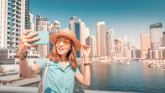 L'image montre une jeune femme prenant un selfie au bord de l'eau, probablement dans une ville dynamique. Elle porte un chapeau en paille et des lunettes de soleil orange, ajoutant une touche estivale à son look. Elle sourit joyeusement, semblant profiter de son moment. En arrière-plan, on peut voir des gratte-ciel modernes et un port avec des bateaux, évoquant un environnement urbain animé et ensoleillé.