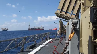تظهر الصورة مشهدًا من سطح سفينة بحرية، حيث يمكن رؤية مجموعة من المنصات البحرية في البحر. المياه تبدو هادئة تحت سماء زرقاء مع بعض السحب البيضاء. من الجانب الأيسر، توجد معدات عسكرية أو دفاعية، ربما صواريخ أو أنظمة توجيه، بينما توجد منصات نفطية في الخلفية. تظهر أيضًا سفن أخرى على مسافة قريبة، مما يدل على نشاط بحري مكثف في المنطقة.