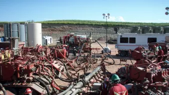 L'image montre un site d'exploitation, probablement lié à l'industrie pétrolière ou gazière. On y voit plusieurs équipements imposants, notamment des pompes et des tuyaux, ainsi que des travailleurs en tenue de sécurité, portant des casques et des vêtements rouges. Le paysage en arrière-plan est rural, avec une colline et des installations industrielles. Le ciel est dégagé, laissant suggérer une journée ensoleillée. L'ensemble de la scène évoque une activité intense et technique, généralement associée à des opérations d'extraction ou de forage.