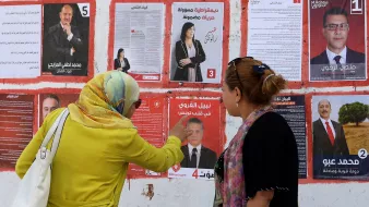 تظهر الصورة جدارًا مغطى بمجموعة من الملصقات الانتخابية. هناك سيدتان تقفان أمام الملصقات. إحداهما ترتدي عباءة ملونة وتظهر وهي تشير إلى أحد الملصقات. يتواجد ملصقات لمرشحين مختلفين، يحمل كل منها صورة واسم المرشح ورقمًا. تعكس الصورة أجواء الانتخابات والحماس المحيط بها، حيث تبدو السيدتان مهتمتين بمناقشة المعلومات المعروضة على الملصقات.
